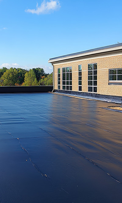 flat black roof surface with a modern building and blue sky in the background features three windows and greenery in the distance